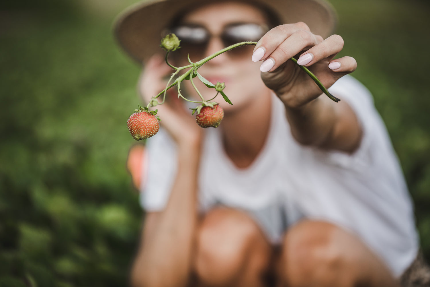 Woman outside with a hat and glasses holding a stem with baby strawberries.